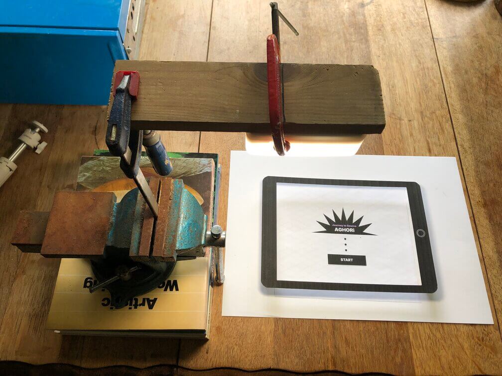 Filming set-up for the paper prototype, using a vice to hold a clamp which holds a piece of wood which has clamped on it a light. My mobile phone then lay down on the plank of wood and filmed the paper prototype underneath.